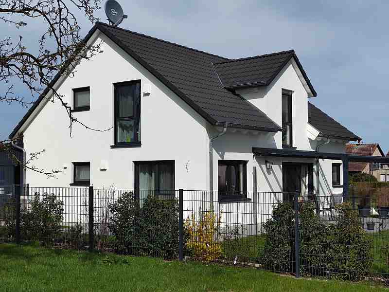 Modernes Einfamilienhaus mit Garten, Zaun und dunklem Satteldach bei Tageslicht.